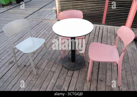 Une scène extérieure minimaliste avec deux chaises roses et une chaise blanche autour d'une table circulaire, sur parquet, créant une ambiance moderne et élégante Banque D'Images