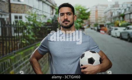 Jeune homme tenant le ballon de football se tient confiant sur la rue urbaine entourée de bâtiments et de voitures, mettant en valeur un look décontracté et sportif. Banque D'Images