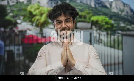 Jeune homme hispanique barbu dans des lunettes souriant avec les mains ensemble dans un geste de prière à l'extérieur entouré de verdure et de montagnes Banque D'Images