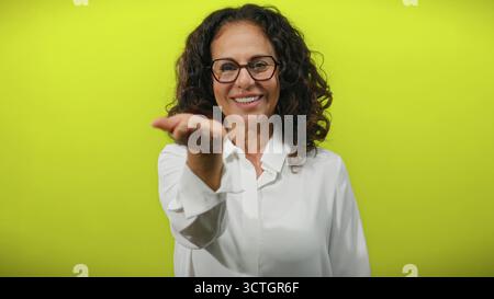 Femme hispanique aux cheveux bouclés étend sa main vers la caméra sur un fond jaune vif, exsudant la confiance et l'accessibilité. Banque D'Images