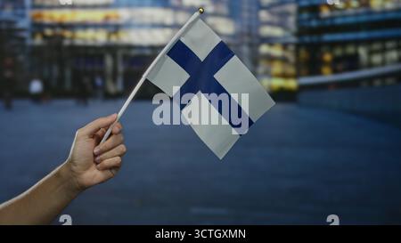 Main tenant le drapeau de la finlande dans la scène de rue de la ville avec fond urbain flou pendant le crépuscule représentant la fierté nationale et la culture. Banque D'Images