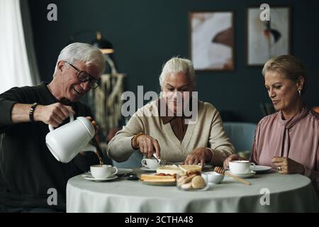 Trois aînés caucasiens assis à la table ronde dégustant le dessert et le café ensemble, un homme senior versant le café tandis que deux amies coupant et servant le gâteau, souriant et interagissant Banque D'Images