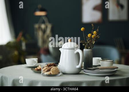Théière en céramique debout sur une table ronde avec des assiettes empilées, des tasses, des biscuits sur un plateau en verre, bouquet de fleurs séchées dans un vase, intérieur de salon moderne floue en arrière-plan Banque D'Images