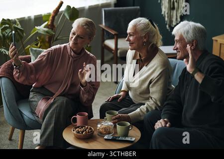 Trois personnes âgées caucasiennes assises ensemble agitant au smartphone pendant un appel vidéo avec des amis, deux femmes et un homme souriant et interagissant, table basse avec des collations et des tasses au premier plan Banque D'Images