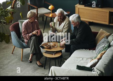Trois femmes caucasiennes âgées et un homme assis ensemble dans le salon buvant des boissons chaudes et parlant, des collations, des biscuits et des biscuits sur la table basse, rassemblement social détendu entre amis Banque D'Images