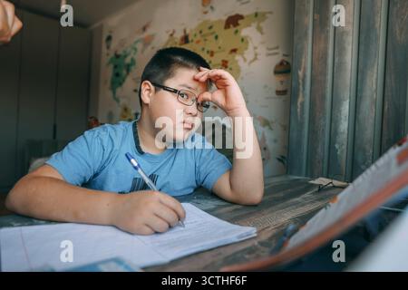 Garçon asiatique avec des lunettes tenant la tête dans la frustration, tâche d'école au bureau à la maison près du fond d'écran de la carte du monde, concept de homeschooling, service d'éducation Banque D'Images