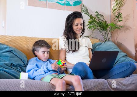 Mère travaillant de la maison sur ordinateur portable tandis que son fils joue tranquillement avec un cube de puzzle coloré Banque D'Images