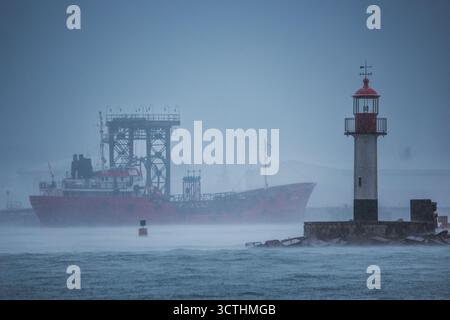 Misty Harbor scène avec Red Cargo Ship et phare sur la côte dans le brouillard et la pluie à Dawn Stormy Weather Varna port maritime, Bulgarie Banque D'Images