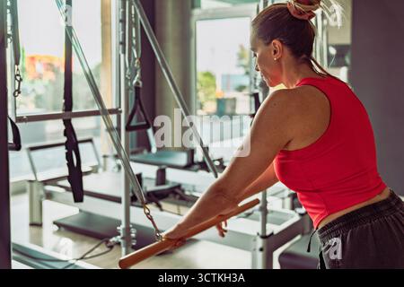 Femme méconnaissable de derrière dans un gros plan détaillé, s'exerçant avec la barre de poussée sur un réformateur Pilates cadillac dans une salle de sport. Banque D'Images