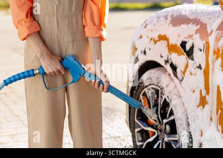 Mains tenant un nettoyeur haute pression à un lave-auto mousseux avec du savon sur la roue et la carrosserie, parfait pour le service d'entretien automobile et les thèmes d'entretien de bricolage Banque D'Images