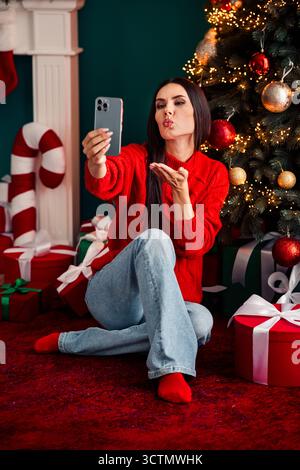 Jeune femme prenant un selfie devant un sapin de Noël tout en célébrant les vacances à la maison dans une ambiance chaleureuse Banque D'Images