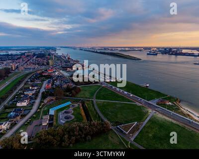 Vue aérienne du port fluvial au coucher du soleil des navires de fret, des grues et des quais industriels bordent les deux rives, avec une grille urbaine dense et des champs verts encadrant l'ouate Banque D'Images