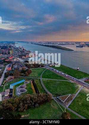 Vue aérienne du port fluvial au coucher du soleil des navires de fret, des grues et des quais industriels bordent les deux rives, avec une grille urbaine dense et des champs verts encadrant l'ouate Banque D'Images
