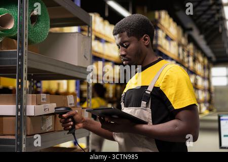 Homme afro-américain utilisant un scanner de codes-barres pour traiter les étiquettes d'expédition sur les boîtes des racks de stockage industriels. Scanner les étiquettes awb pour le service de suivi des colis avec des informations de suivi, une logistique précise. Banque D'Images