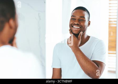 Joyeux jeune Noir Homme regardant dans le miroir dans la salle de bains et face touchante Banque D'Images