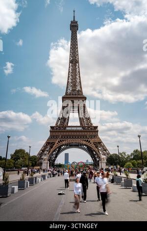 Foules marchant près de la Tour Eiffel à Paris sous un ciel lumineux d'été avec des anneaux olympiques affichés. Banque D'Images