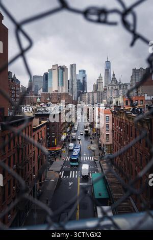 Vue sur le paysage urbain à travers une clôture à maillons de chaîne, avec une rue animée avec des voitures et de hauts gratte-ciel en arrière-plan, sous un ciel nuageux. Banque D'Images
