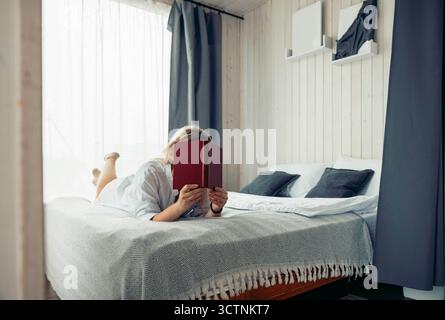 Une jeune femme caucasienne aux cheveux blonds est allongée sur un lit, lisant un livre. La chambre dispose de rideaux lumineux et d'une atmosphère chaleureuse, idéale pour une escapade de vacances. Banque D'Images