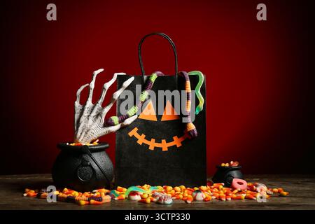 Chaudrons, sac en papier, main squelette et délicieux bonbons à la gelée d'Halloween sur la table en bois près du mur rouge Banque D'Images