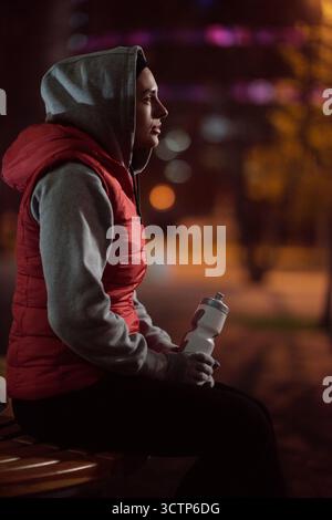 Femme se reposant après l'entraînement. Vue latérale de la coureuse féminine assise sur le banc avec bouteille de sport le soir d'automne et regardant dans la distance sur fond flou Banque D'Images