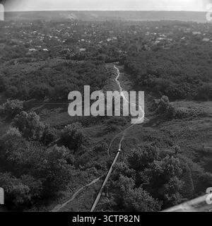 Une vue panoramique sur la région de 'Vinogradniki' (vignobles) dans le microdistrict Magnolia de Sloviansk, RSS d'Ukraine, au cours des années 1970 Cette photographie d'archives capture une partie remarquablement verte, presque rurale de la ville. Le paysage est dominé par des jardins luxuriants, des vignobles privés et la canopée d'arbres entourant les maisons d'un étage. Un pipeline industriel traverse le paysage. Au loin, un lac offre une séparation naturelle des parties plus urbaines de la ville visibles à l'horizon. Banque D'Images