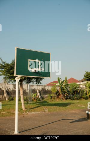 Un panneau de basket-ball vert foncé altéré se dresse contre un ciel bleu clair et un mur de béton brisé dans un terrain extérieur ensoleillé et négligé Banque D'Images