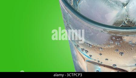 Gros plan d'un verre transparent rempli de glace et d'une boisson gazeuse. Présence de condensation et de bulles visibles sur un fond vert éclatant. Banque D'Images