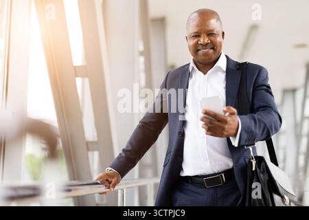 Heureux homme d'affaires afro-américain utilisant smartphone et souriant dans l'aéroport moderne Banque D'Images
