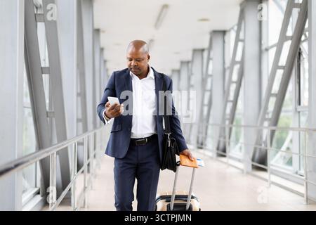 Homme d'affaires noir marchant dans l'aéroport, vérifiant smartphone et tenant passeport avec valise Banque D'Images