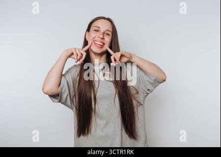 Jeune femme souriant et pointant ses joues sur un fond gris. Banque D'Images