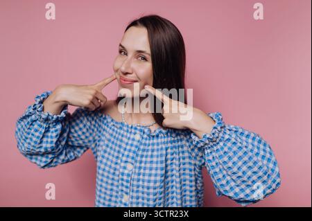 Femme souriante en robe bleue à carreaux pointant vers ses joues. Banque D'Images