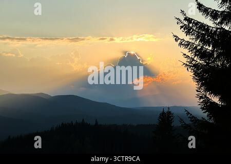 Coucher de soleil spectaculaire sur les collines, avec les rayons du soleil perçant à travers un nuage unique, projetant une ombre étonnante. Beauté sereine de la nature avec un ciel vif et des couches de montagnes bleues en arrière-plan. Banque D'Images