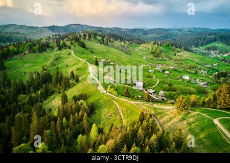 Photographie aérienne du paysage rural serein en Ukraine, avec des collines verdoyantes, des chemins de terre sinueux et des maisons dispersées. Des forêts denses entourent la zone, avec un ciel spectaculaire et nuageux au-dessus. Banque D'Images