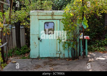 Ancien hangar turquoise avec peinture pelable, porte cadenassée, et cadre rustique dans un jardin verdoyant, évoquant le charme de la campagne vintage Banque D'Images