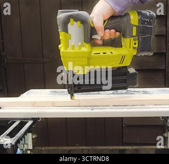 L'ouvrier pousse des clous dans la poutre en bois à l'aide d'un pistolet à clous alimenté par batterie sur le chantier sous le jour des travaux Banque D'Images