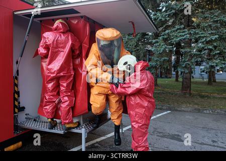 Les membres de l'équipe d'urgence portant des combinaisons chimiques rouges aident un troisième intervenant portant une combinaison de matières dangereuses orange à pénétrer dans une unité de décontamination mobile ou dans un véhicule. Banque D'Images