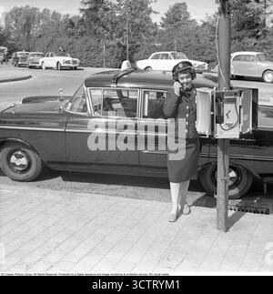Chauffeur de taxi féminin dans les années 1960 Dans les années 1950 et 1960, les chauffeuses de taxi étaient une présence rare mais frappante sur les routes. Défiant les normes de genre, ces femmes ont navigué dans des villes animées, offrant un service vital avec confiance et compétence. Dans les années 1950 et 1960, les téléphones de station de taxis, montés sur des poteaux où les taxis attendaient, permettaient aux chauffeurs de recevoir des réservations directement d'une répartition centrale. Les clients pouvaient appeler le numéro du rang, et la cloche du téléphone alerterait les conducteurs, assurant des réponses rapides dans les zones urbaines achalandées. Suède 1962 Banque D'Images