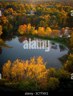 Vue aérienne de la lumière dorée de l'automne reflétant les eaux calmes d'un étang niché parmi les arbres vibrants dans un paysage pittoresque, Easthampton, Massachusetts, États-Unis. Banque D'Images