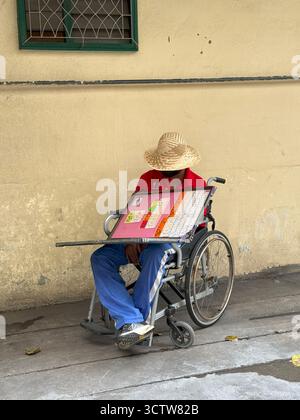 Bangkok, Thaïlande - 27 septembre 2025 : un homme en fauteuil roulant portant un chapeau de paille et une chemise rouge est assis près d'un mur beige, vendant des billets de loterie sur le s. Banque D'Images