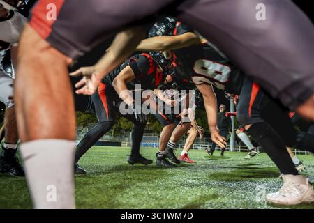 Joueurs de football américain accroupis sur un gazon artificiel vert sous les lumières du stade, alignés pour un scrimmage de nuit, concentrés et prêts pour une équipe intense Banque D'Images