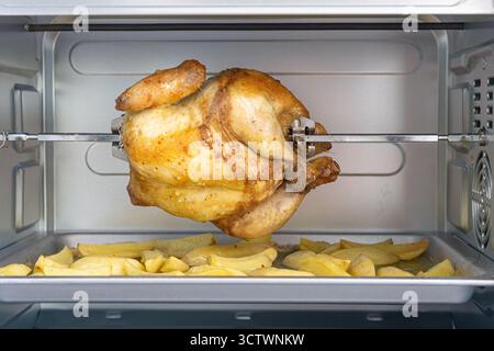 Poulet rôti doré et juteux sur une broche au-dessus d'un plateau de quartiers de pommes de terre assaisonnés dans un four chaud et maison, invitant des ambiances réconfortantes pour le dîner en famille. Banque D'Images