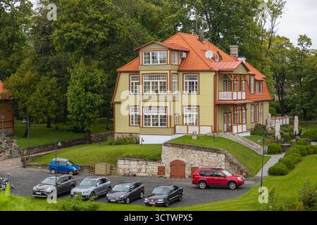 Kuressaare, Saaremaa, Estonie – 25 août 2021 : Villa en bois restaurée avec toit orange et voitures garées devant, entourée d'arbres verts. Banque D'Images