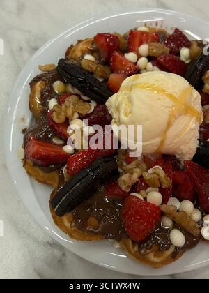 L'image montre un gros plan d'une assiette à dessert avec une gaufre recouverte de pâte à tartiner au chocolat, de fraises fraîches, de biscuits Oreo, de noix et de chocolat blanc Banque D'Images