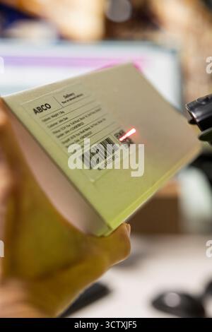 Gros plan sur un employé asiatique qui lit des étiquettes de suivi et des codes-barres sur les colis, vérifiant l'inventaire des marchandises avec un scanner. Systèmes de centre de distribution gérant une livraison express efficace. Banque D'Images