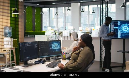 Informaticien dans un bureau de démarrage examinant les métriques du système d'IA, surveillant les tableaux de bord de performance. Femme afro-américaine accordant des réseaux neuronaux en utilisant des outils sur PC, caméra A. Banque D'Images