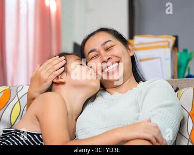 Une fille asiatique aimante embrassant sa mère souriante sur la joue tout en la serrant sur le canapé à la maison. Banque D'Images