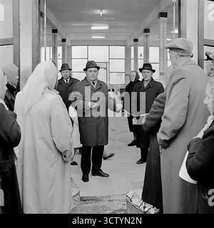 Une photo vintage en noir et blanc de 1970 capture la grande ouverture d'un nouveau marché couvert à Sloviansk, en RSS d'Ukraine. Un homme portant un chapeau et un manteau, probablement un officiel, coupe cérémoniellement un ruban, symbolisant l'inauguration. Une foule de spectateurs enthousiastes et de premiers acheteurs, dont beaucoup ont des paniers vides, se rassemble autour. L'intérieur lumineux et moderne avec de grandes fenêtres et colonnes reflète un sentiment de progrès. Cet événement a été un jalon important pour les infrastructures urbaines, mettant en évidence les améliorations dans la vie quotidienne en URSS, particulièrement significatives lors du 100e anniversaire de la naissance de Lénine Banque D'Images