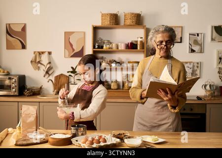 Femme âgée caucasienne lisant un livre de recettes tandis qu'une fille caucasienne mélangeant des ingrédients dans un bol en verre sur le comptoir de la cuisine, portant des tabliers et préparant la nourriture ensemble à l'intérieur Banque D'Images