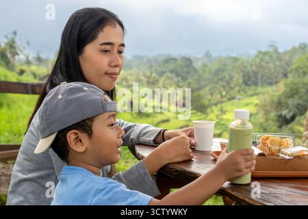 Une mère souriant à son tout-petit tout en tenant une bouteille de boisson à une table en bois près des rizières vertes. Banque D'Images