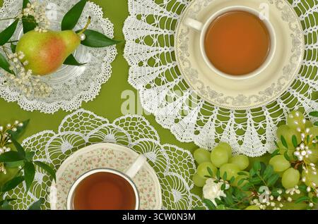 Thé, poires, raisins, perles et fleurs sur doilies crochetées et fond vert. Vue de dessus table vintage dans le salon de thé. Banque D'Images